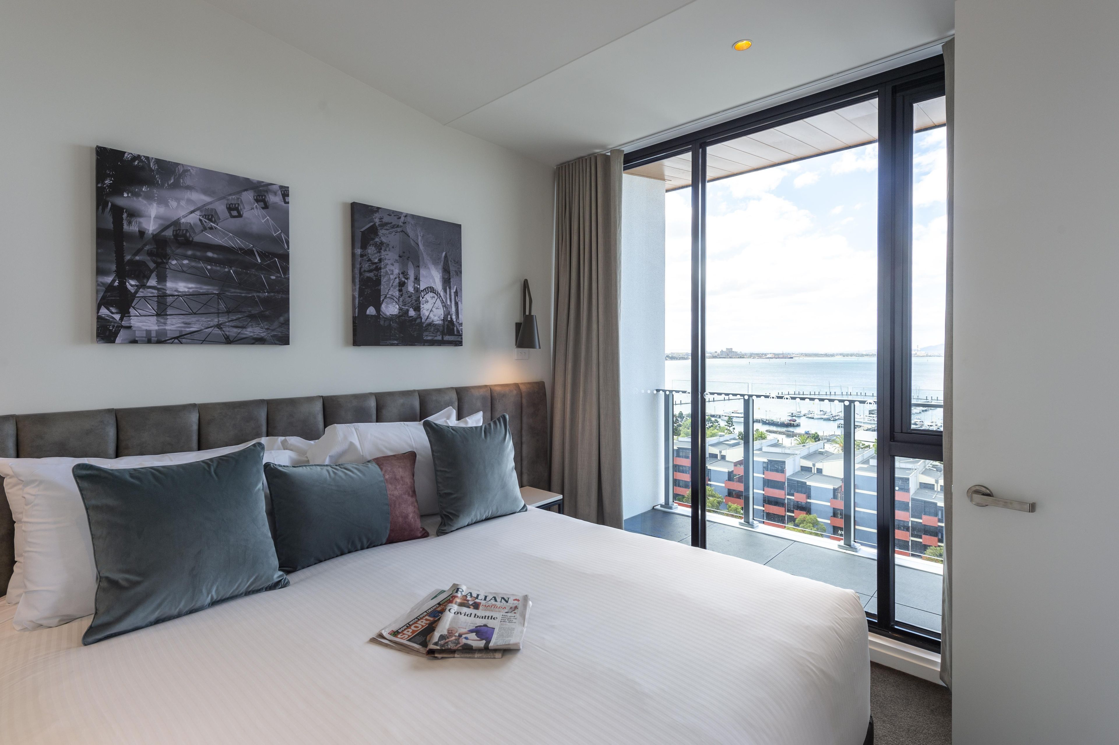 Une chambre d’hôtel avec un grand lit, deux œuvres d’art accrochées au mur, une fenêtre donnant sur la ville et un balcon avec une balustrade.