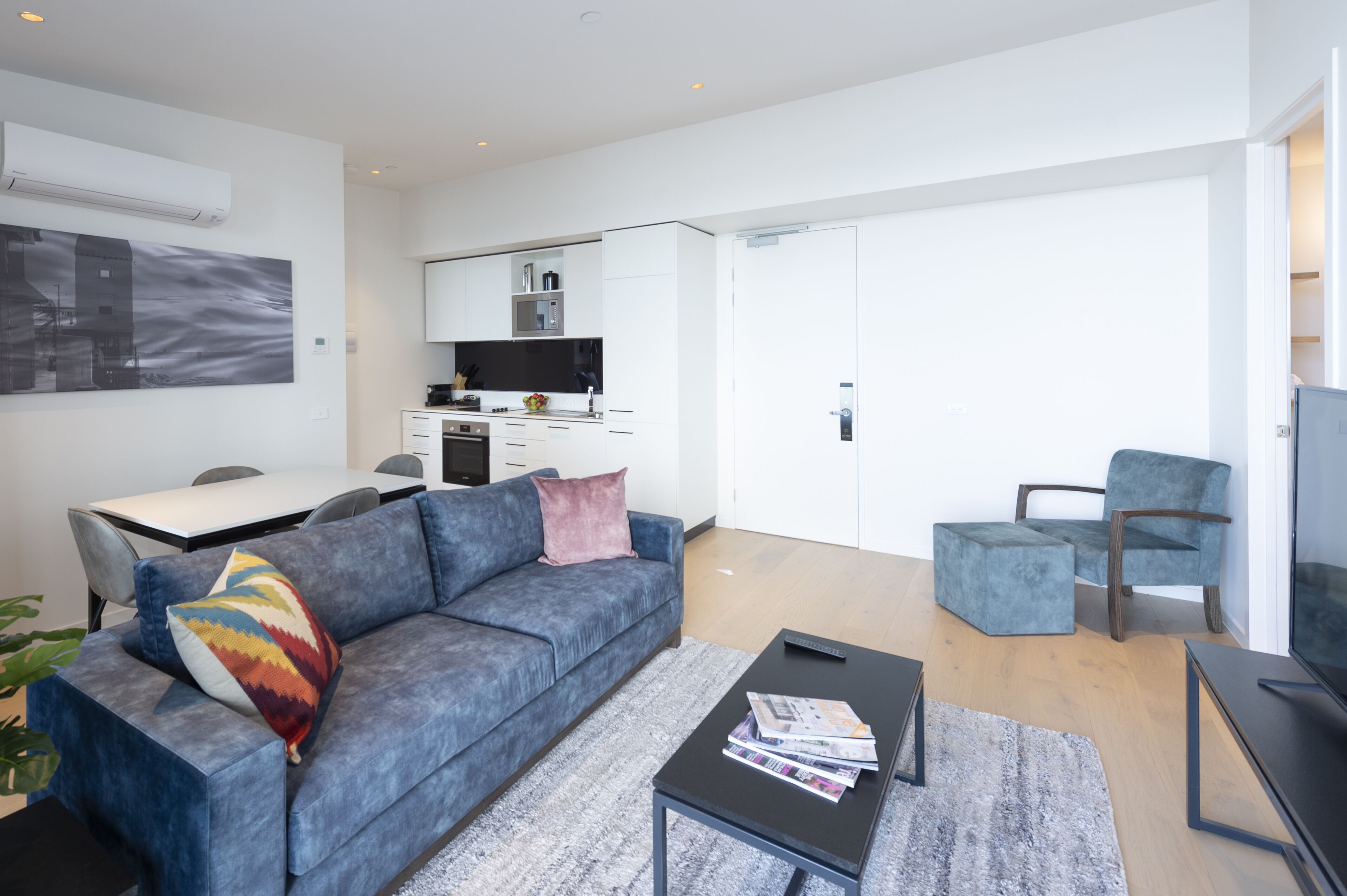 Een moderne woonkamer met een grijze bank, een salontafel en een televisie. Er is een eethoek met een tafel en stoelen, en een keuken met witte kastjes en een magnetron.