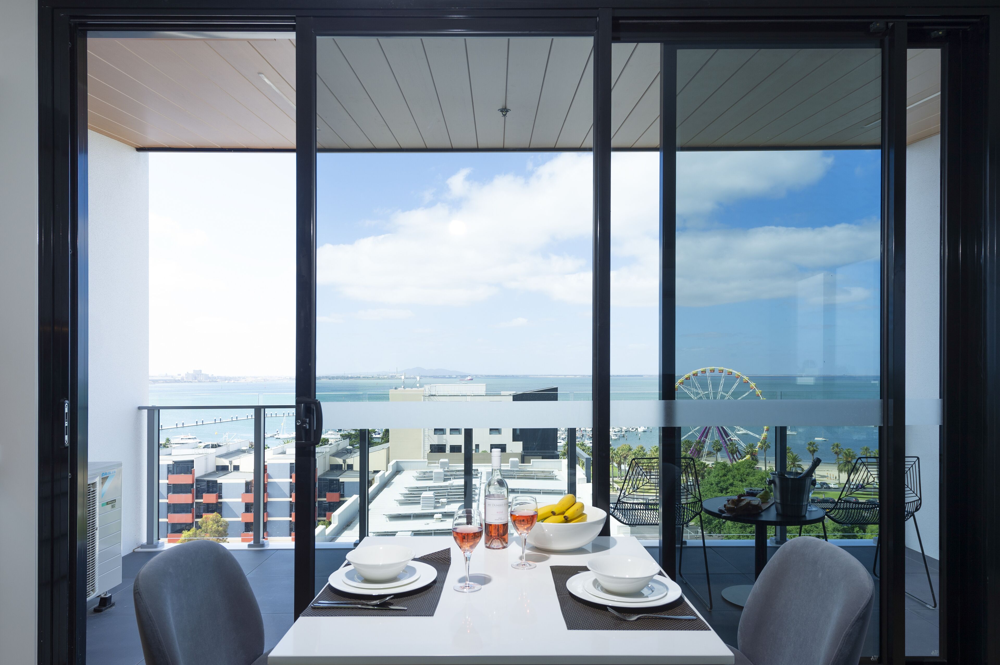 A balcony with a table set for two, a bottle of wine, and a bowl of fruit.