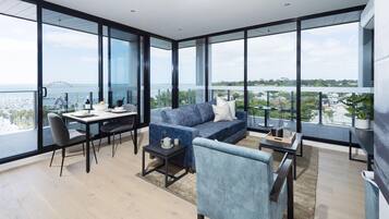 A modern living room with a sofa, a dining table, and chairs, large windows offering a view of the sea and a cityscape.