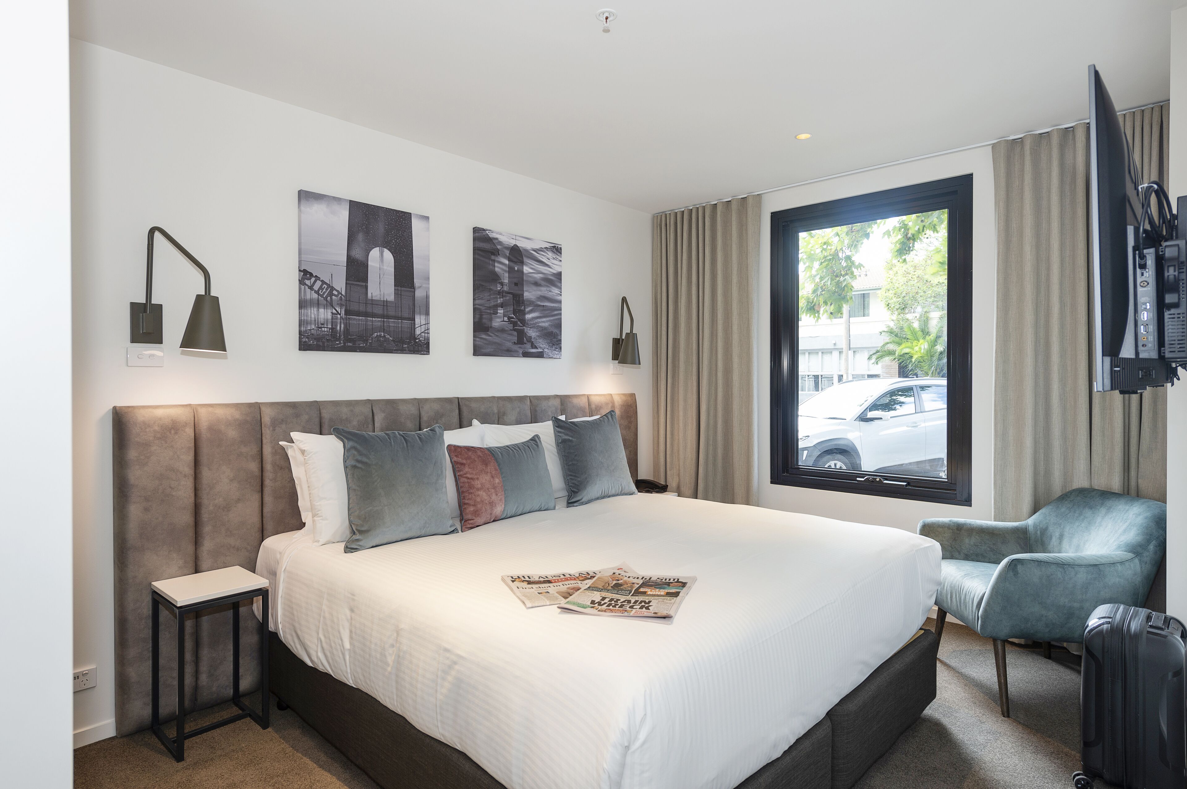 A hotel room with a large bed, a small table, a chair, a TV, and a window with a view of the street.