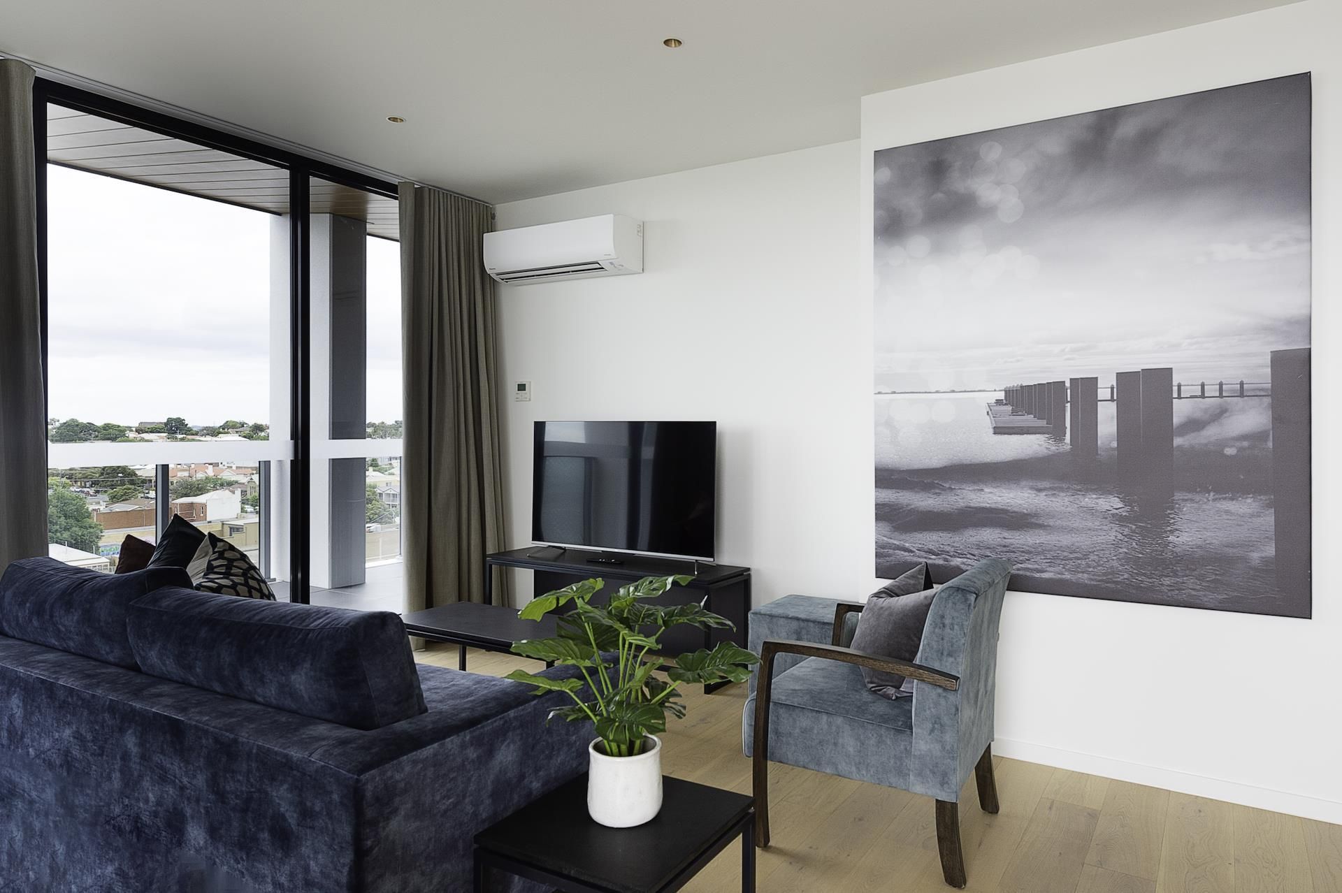 A modern living room with a large window, a flat-screen TV, a grey sofa, a small table with a plant, and a large black-and-white photograph on the wall.