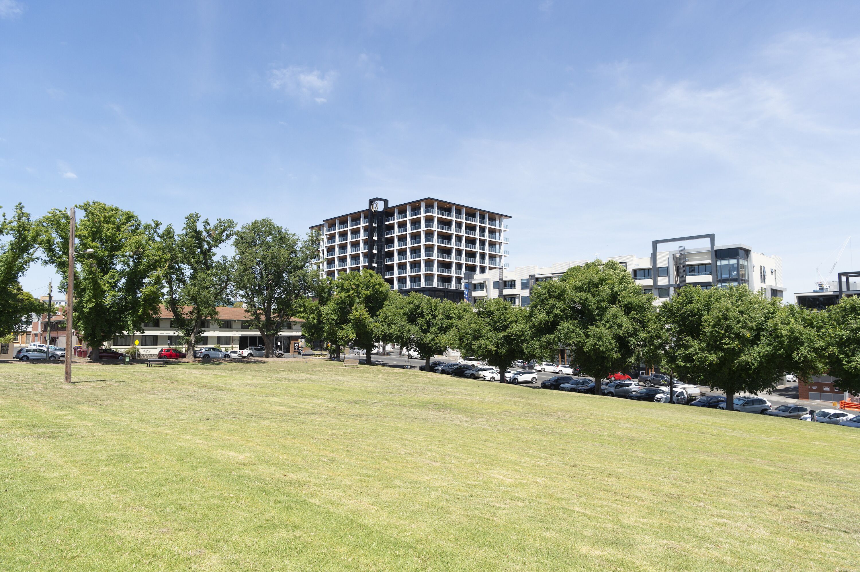 Un campo cubierto de hierba con árboles y coches estacionados, un edificio moderno de varios pisos, y otro edificio contemporáneo con un diseño singular.