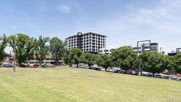 Un campo cubierto de hierba con árboles y coches estacionados, un edificio moderno de varios pisos, y otro edificio contemporáneo con un diseño singular.