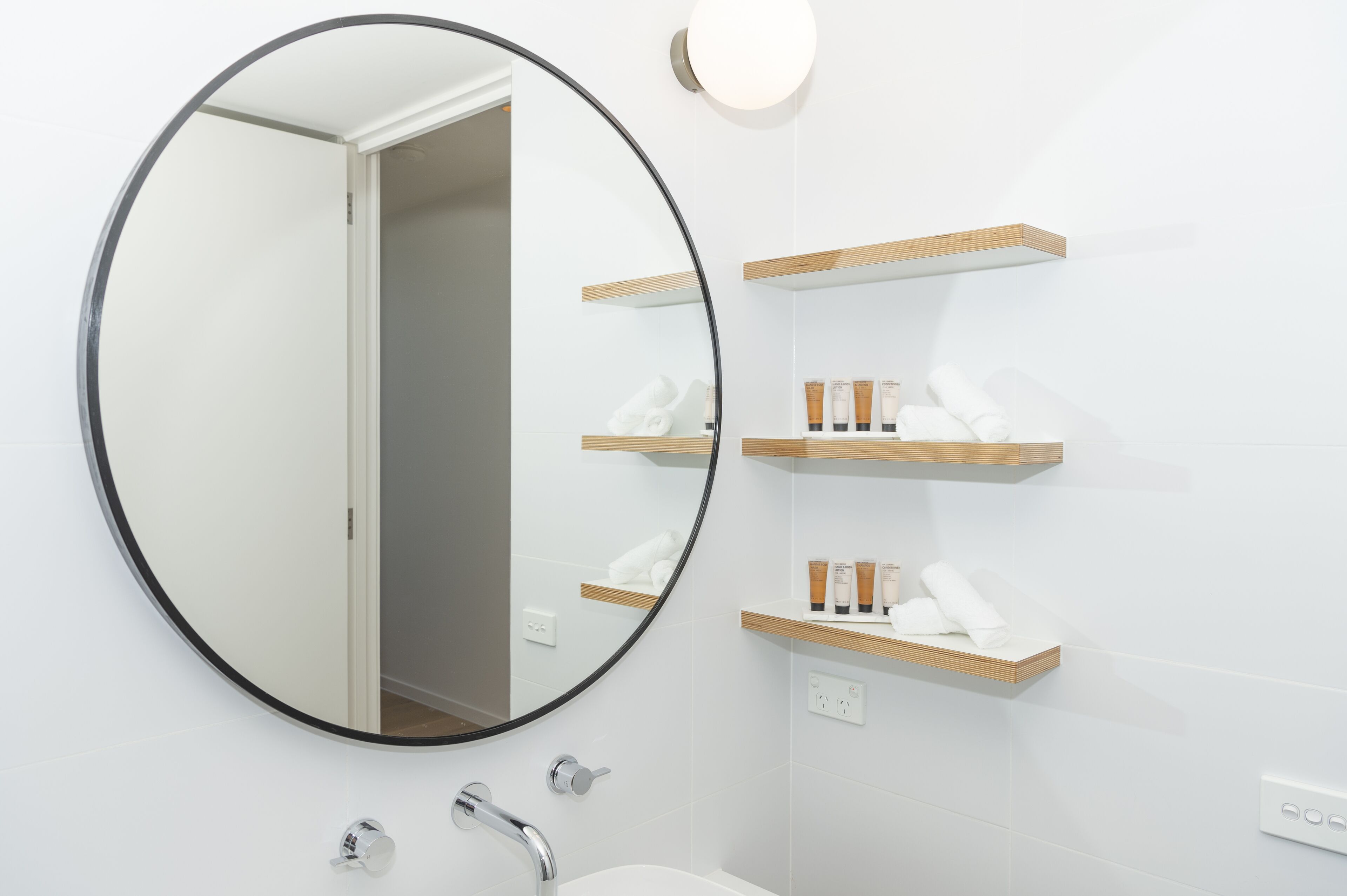 A bathroom with a round mirror, a sink, and wooden shelves with toiletries.