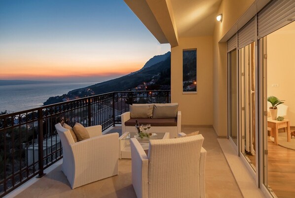 A balcony with a view of the sea at sunset, furnished with white wicker chairs and a table, and a sliding glass door leading to an indoor area with a plant.