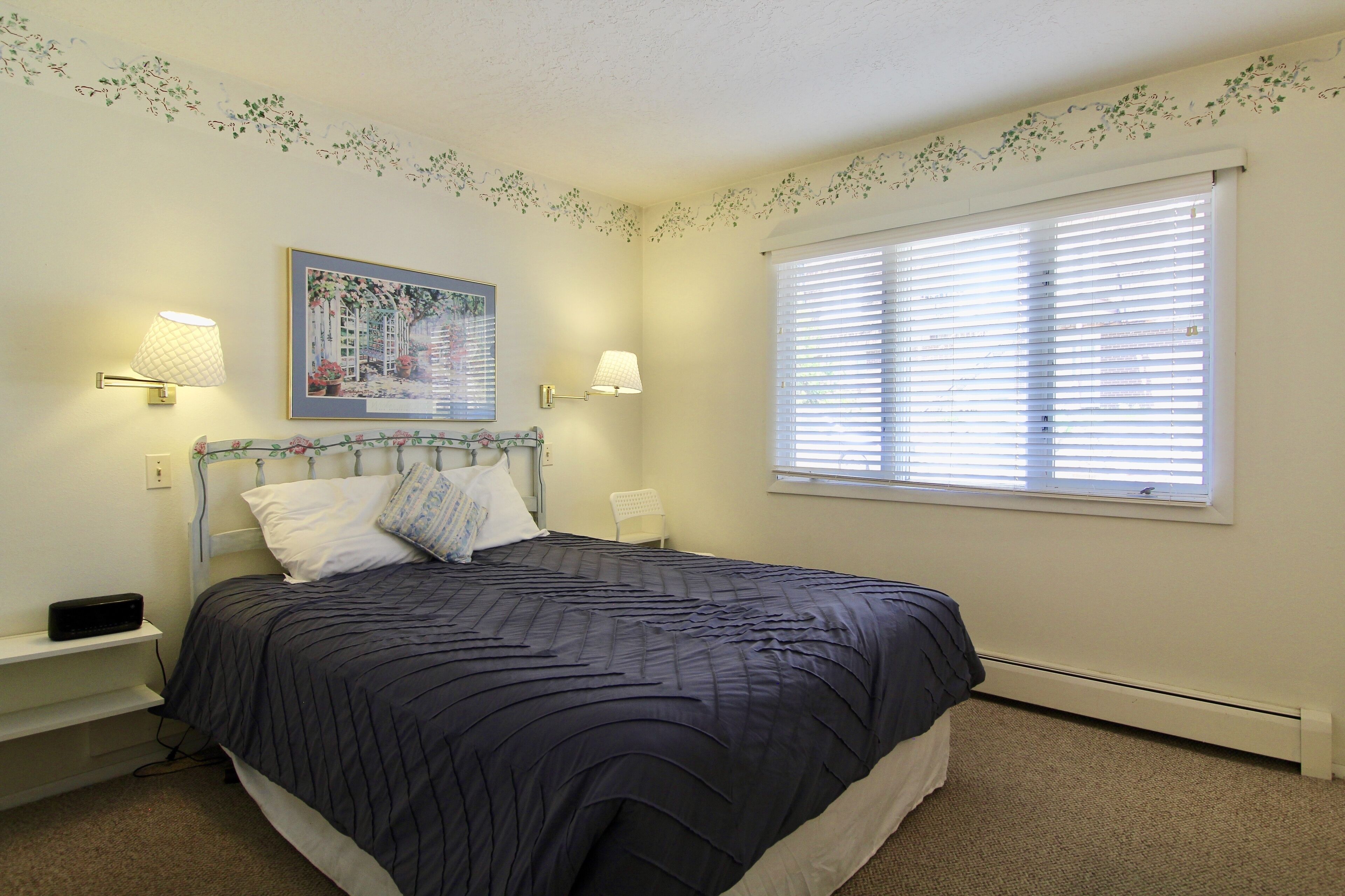 A bedroom with a bed, two bedside lamps, a nightstand, a window with blinds, and a wall-mounted picture.