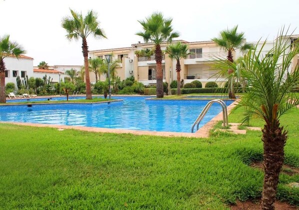 A swimming pool surrounded by palm trees and a grassy area, with buildings in the background.