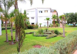 A modern building with a white facade, surrounded by a well-maintained garden with various shrubs and palm trees.
