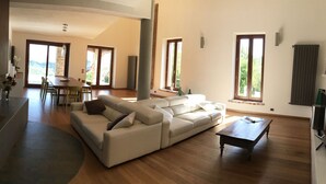 A spacious living area with a large L-shaped sofa, a dining table, and a coffee table. There are multiple windows providing natural light and views of greenery outside.