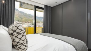Quarto de hotel moderno com uma cama grande, vista para a cidade e montanhas através de uma varanda, e uma janela ampla com cortinas escuras.