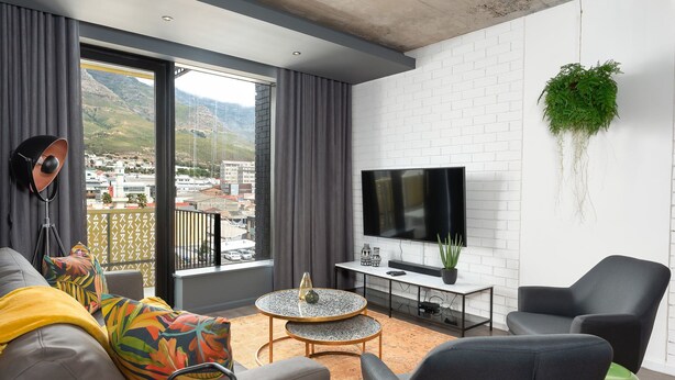 Uma sala de estar moderna com um sofá, cadeiras, uma TV e uma varanda com vista para a cidade e para as montanhas.