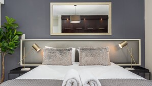 Un lit bien fait, avec deux oreillers moelleux, une couverture tricotée grise et deux serviettes enroulées. On y trouve des tables de chevet avec des lampes, un grand miroir au-dessus du lit et une plante en pot sur le côté gauche.