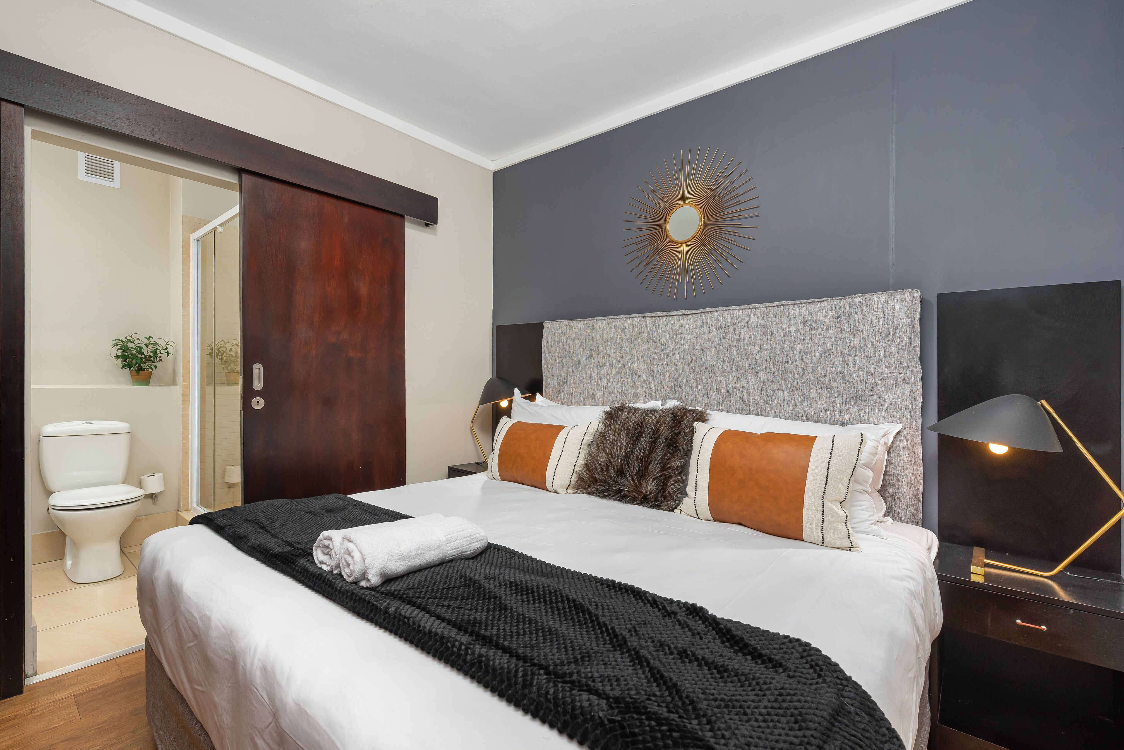 A modern hotel room with a large bed, a dark headboard, a sunburst wall decoration, a bedside lamp, and a view of the bathroom.