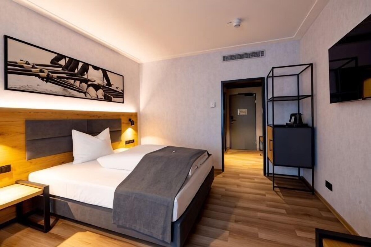 A modern hotel room with a wooden headboard, a bed with white linens, a bedside table, a wall-mounted TV, and a shelf with a coffee machine.