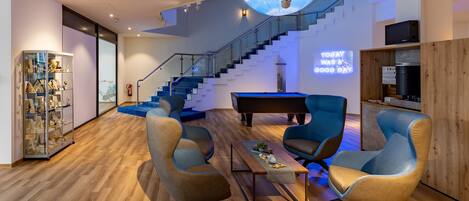 A modern living room with a wooden floor, a grand piano, blue armchairs, and a staircase with a glass railing.
