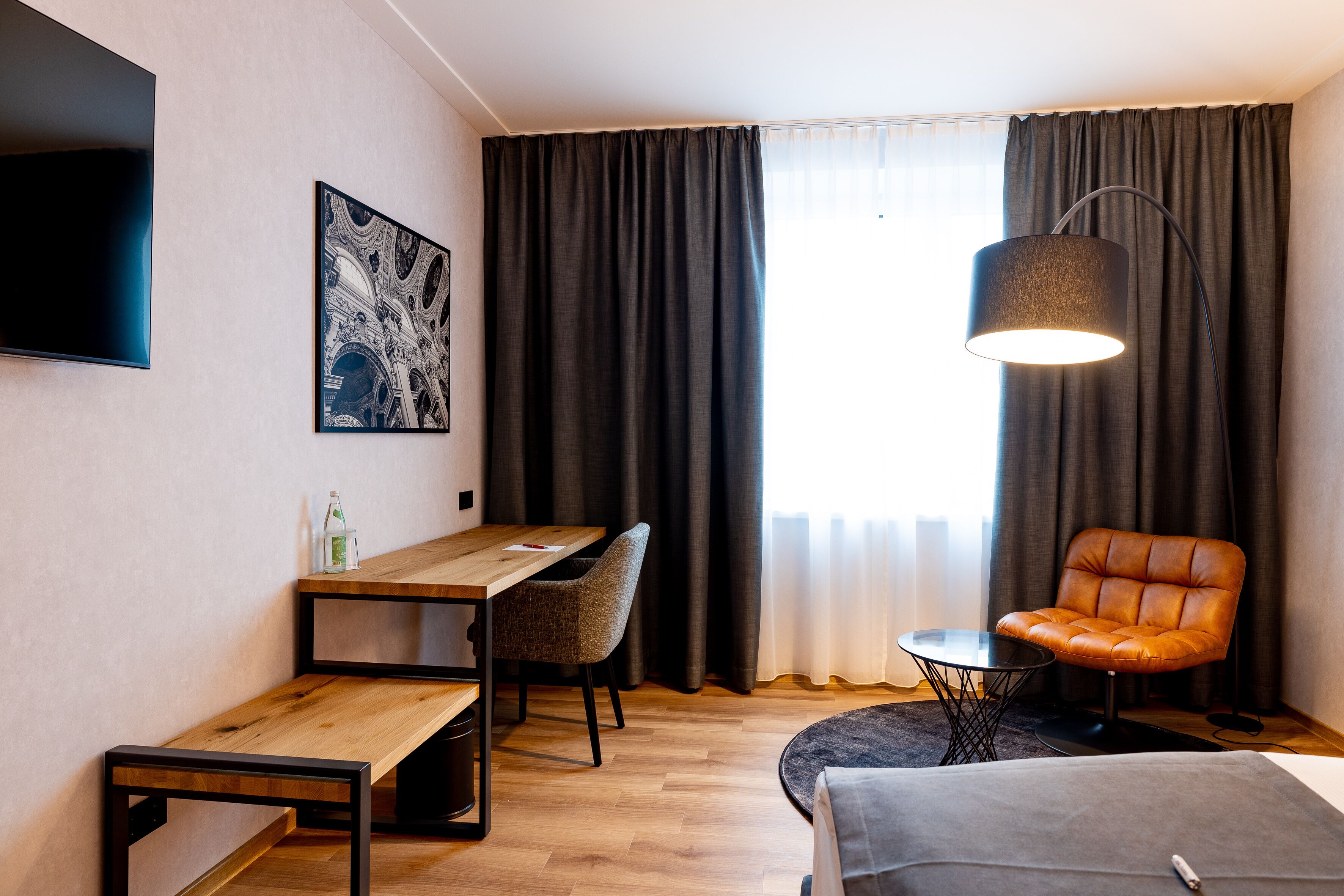 Et moderne hotelværelse med et træskrivebord, en behagelig lænestol, et lille rundt bord og et stort vindue med gardiner.