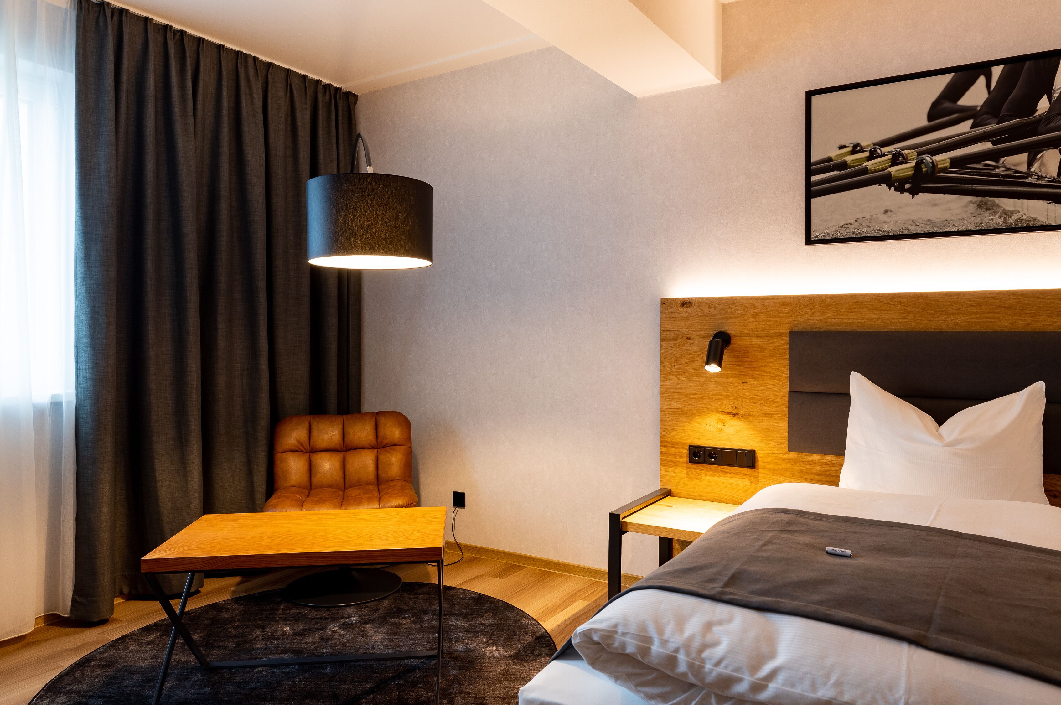 A modern hotel room with a large bed, a leather armchair, a small table, and a wall-mounted lamp.