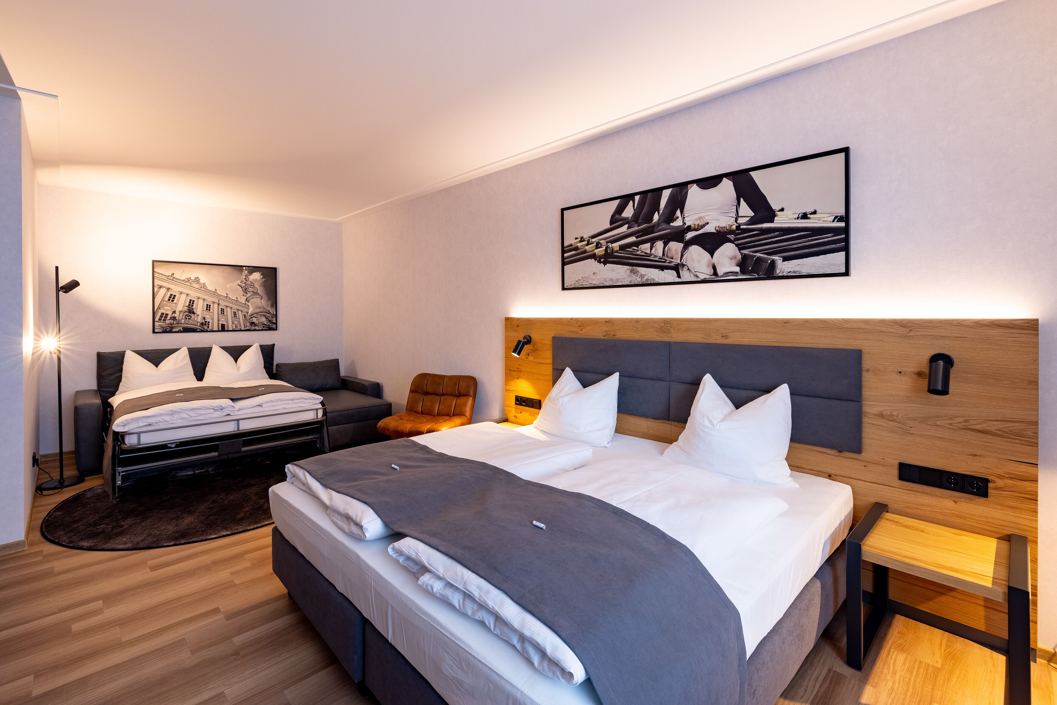 A modern hotel room with a large bed, a sofa, a desk, and a chair. There are two paintings on the wall and a lamp on the floor.