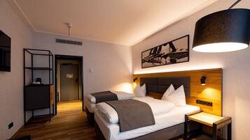 Habitación de hotel con dos camas, piso de madera, un escritorio, una estantería y una lámpara fijada a la pared.