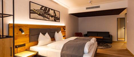 Un dormitorio moderno con cabecera de madera, cama con ropa de cama blanca, mesita de noche, escritorio pequeño, silla, sofá y un cuadro enmarcado en la pared.