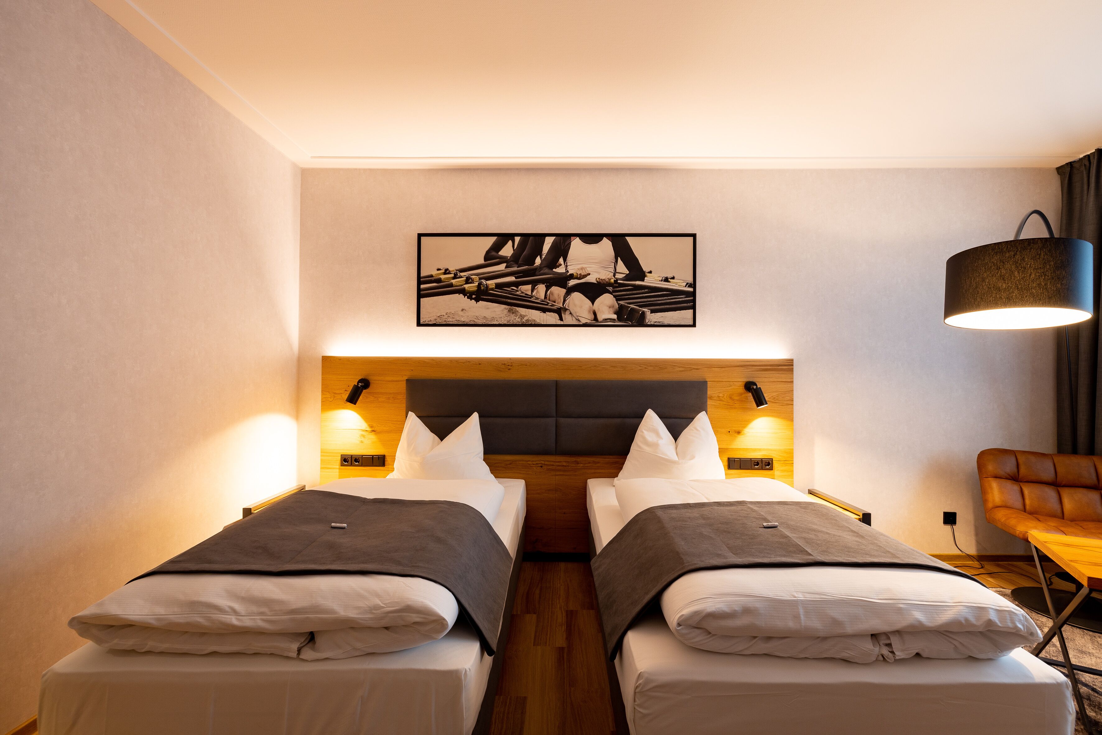 A hotel room with two beds, a wooden headboard, wall-mounted lamps, a framed picture above the beds, and a leather chair.