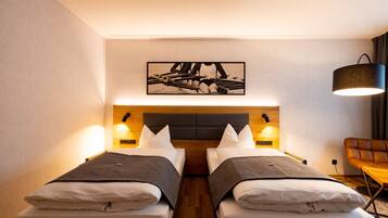 A hotel room with two beds, a wooden headboard, wall-mounted lamps, a framed picture above the beds, and a leather chair.