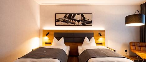 A hotel room with two beds, a wooden headboard, wall-mounted lamps, a framed picture above the beds, and a leather chair.