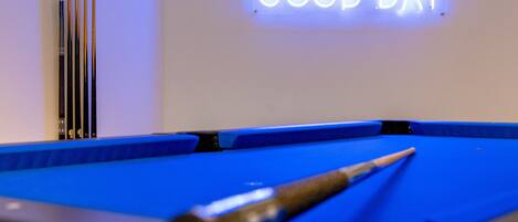 A pool table with a cue stick, a neon sign stating "TOP PAY WAS A GOOD DAY", and a row of pool cues against a white wall.