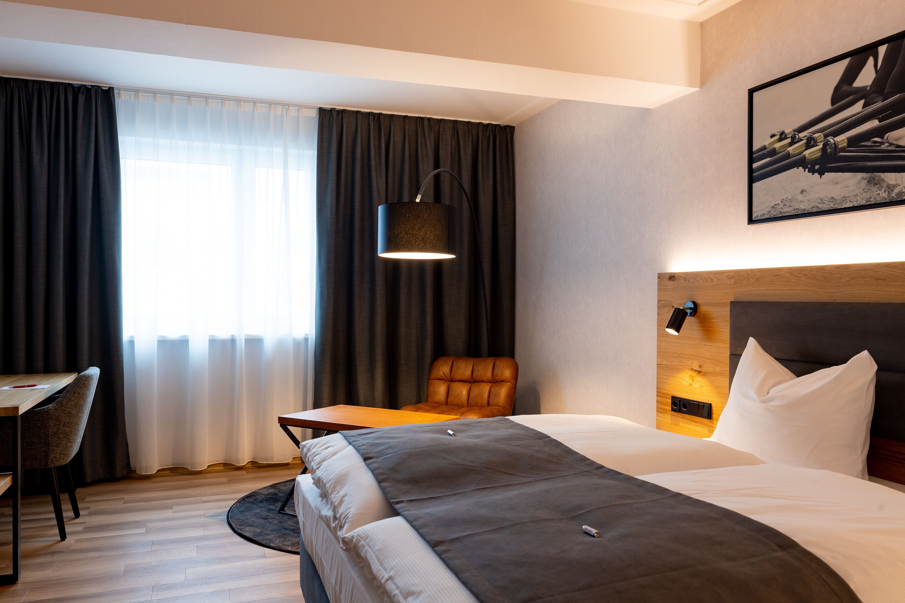 A hotel room with a bed, a desk, a chair, a lamp, and a painting on the wall.
