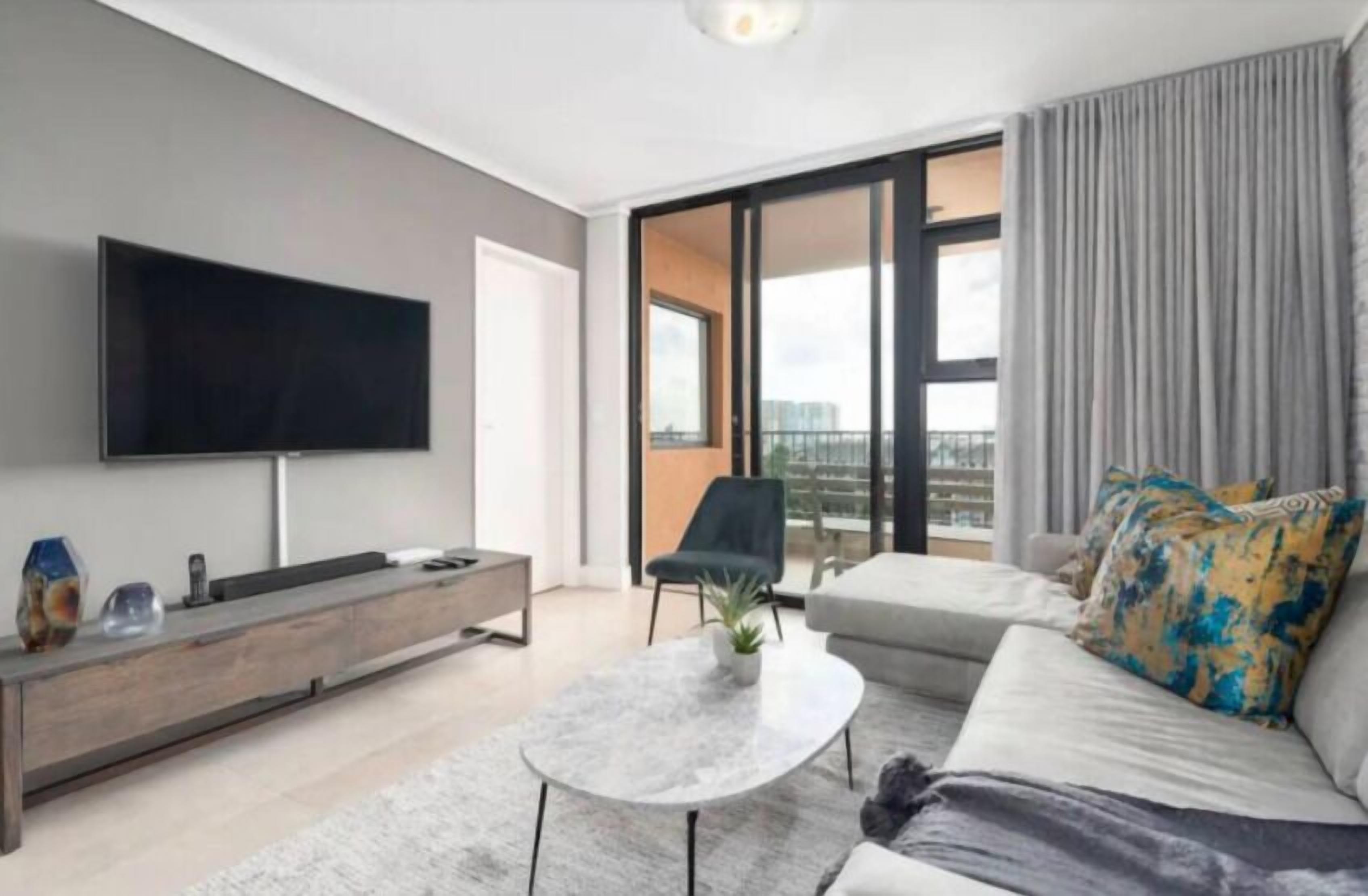 A modern living room with a flat-screen TV, a sofa, a coffee table, and a balcony with curtains.