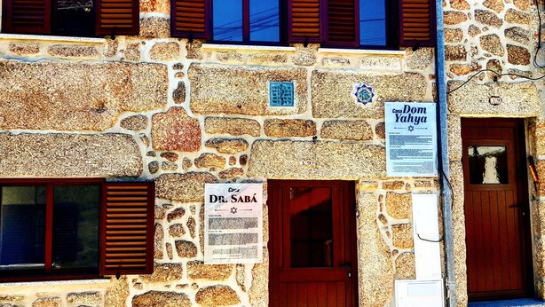 A stone building with multiple wooden shutters, a sign for Casa Dom Yakhya, and a sign for Casa Dr. Sabá. - Casa Dr. Saba (Belmonte, Portugal)