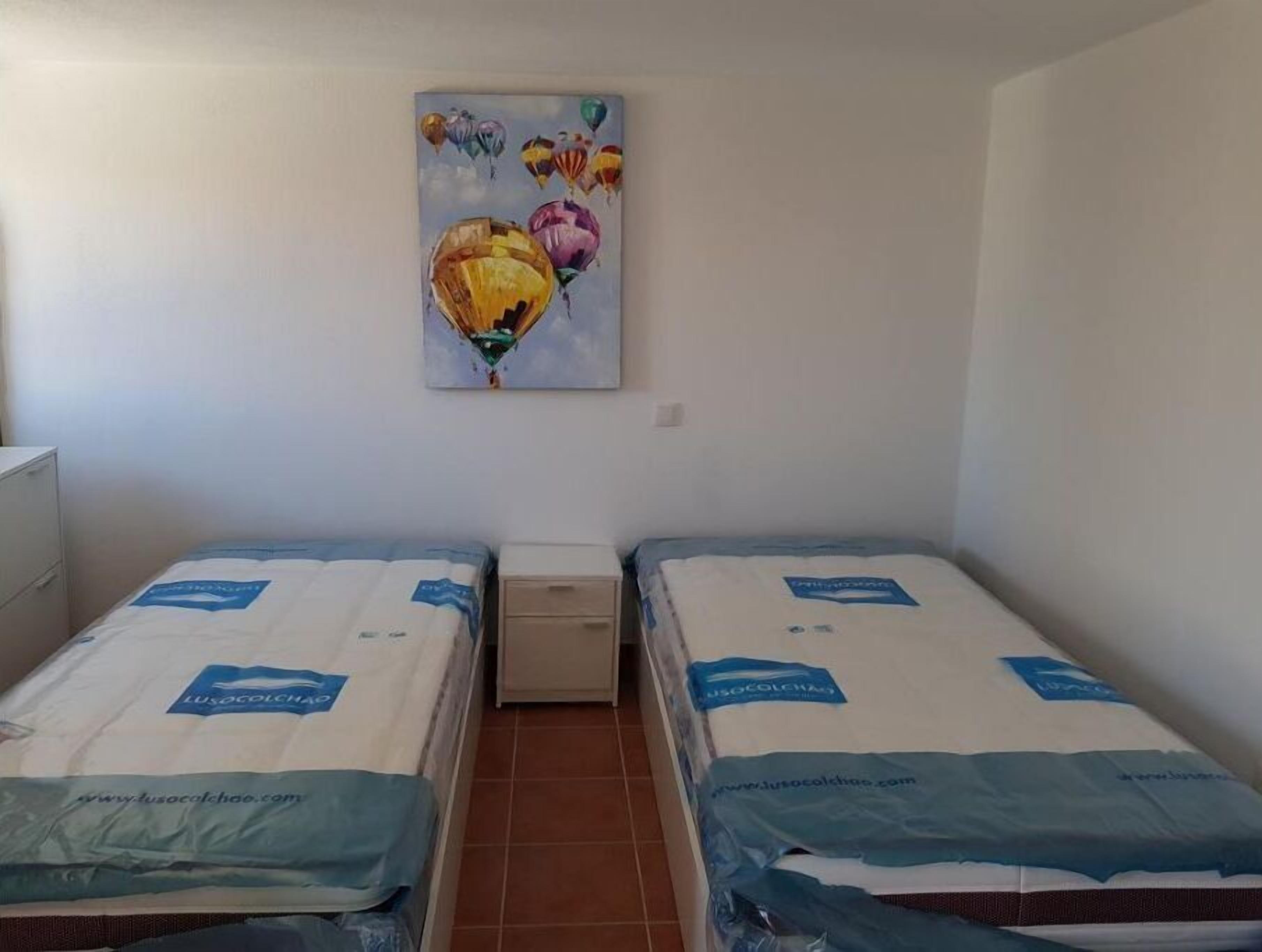 Quarto pequeno e limpo com duas camas de solteiro, cada uma coberta por um colchão branco e um cobertor azul. Um quadro com balões coloridos está pendurado na parede acima das camas.