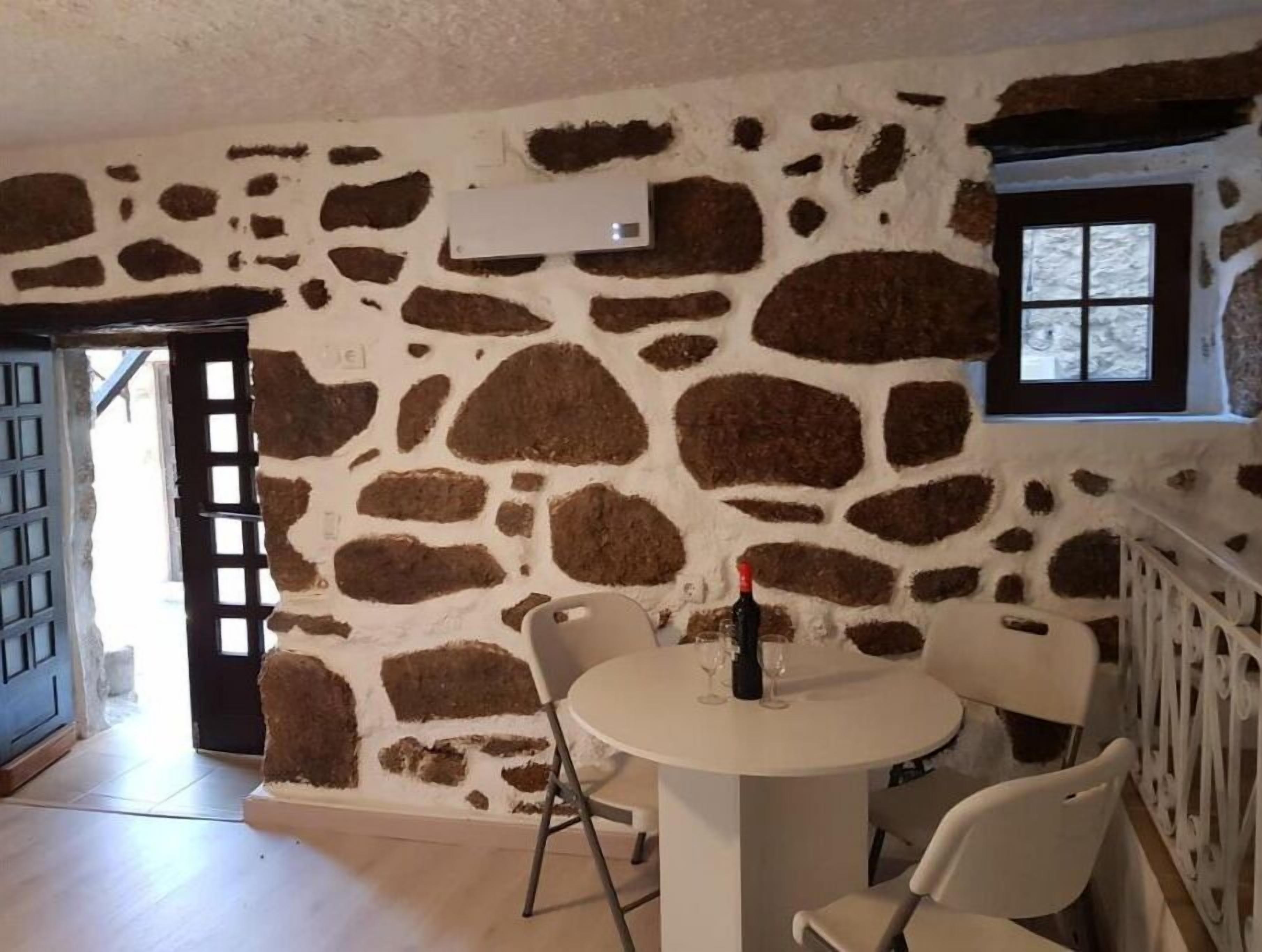 A room with a stone wall, a round table with chairs, a bottle of wine, and a window with a view.