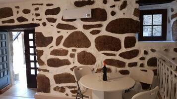 A room with a stone wall, a round table with chairs, a bottle of wine, and a window with a view.