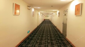 Un pasillo largo y estrecho de hotel con alfombra estampada, cuadros en las paredes y un detector de incendios a la izquierda.
