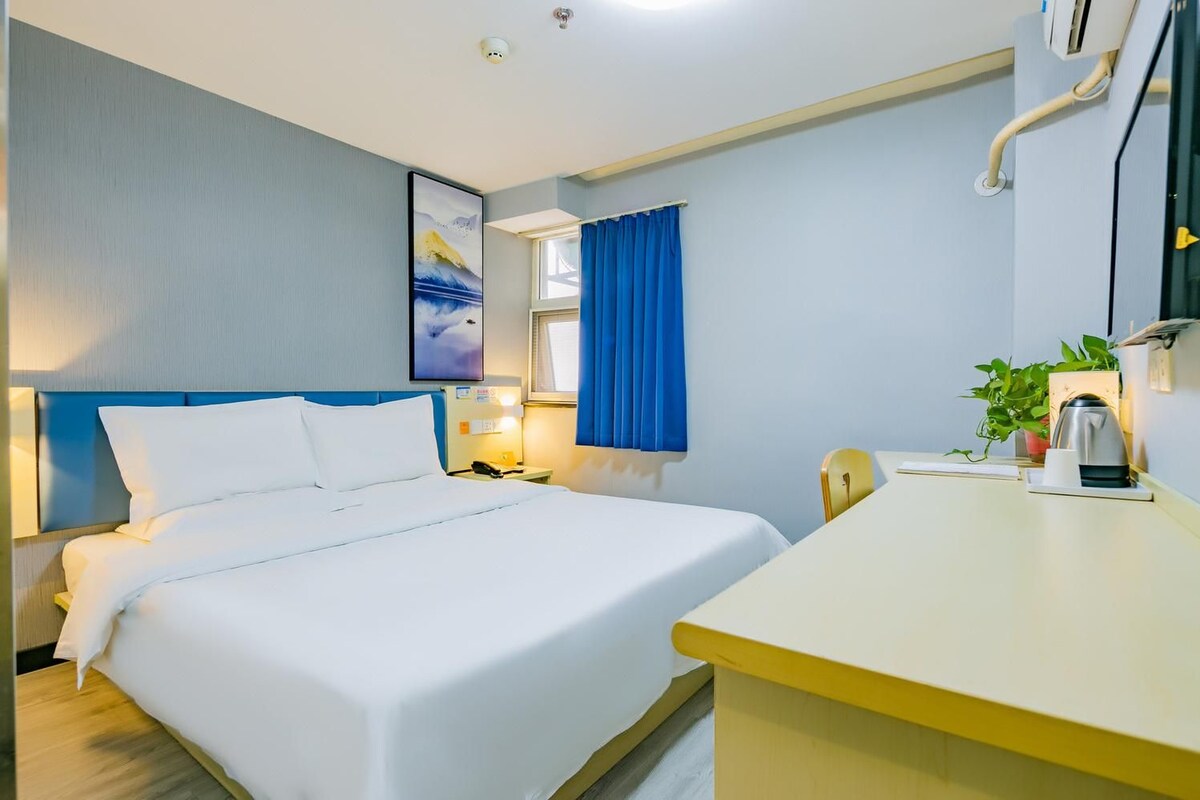 A hotel room with a bed, bedside table, lamp, window with curtains, and a small desk with a kettle.