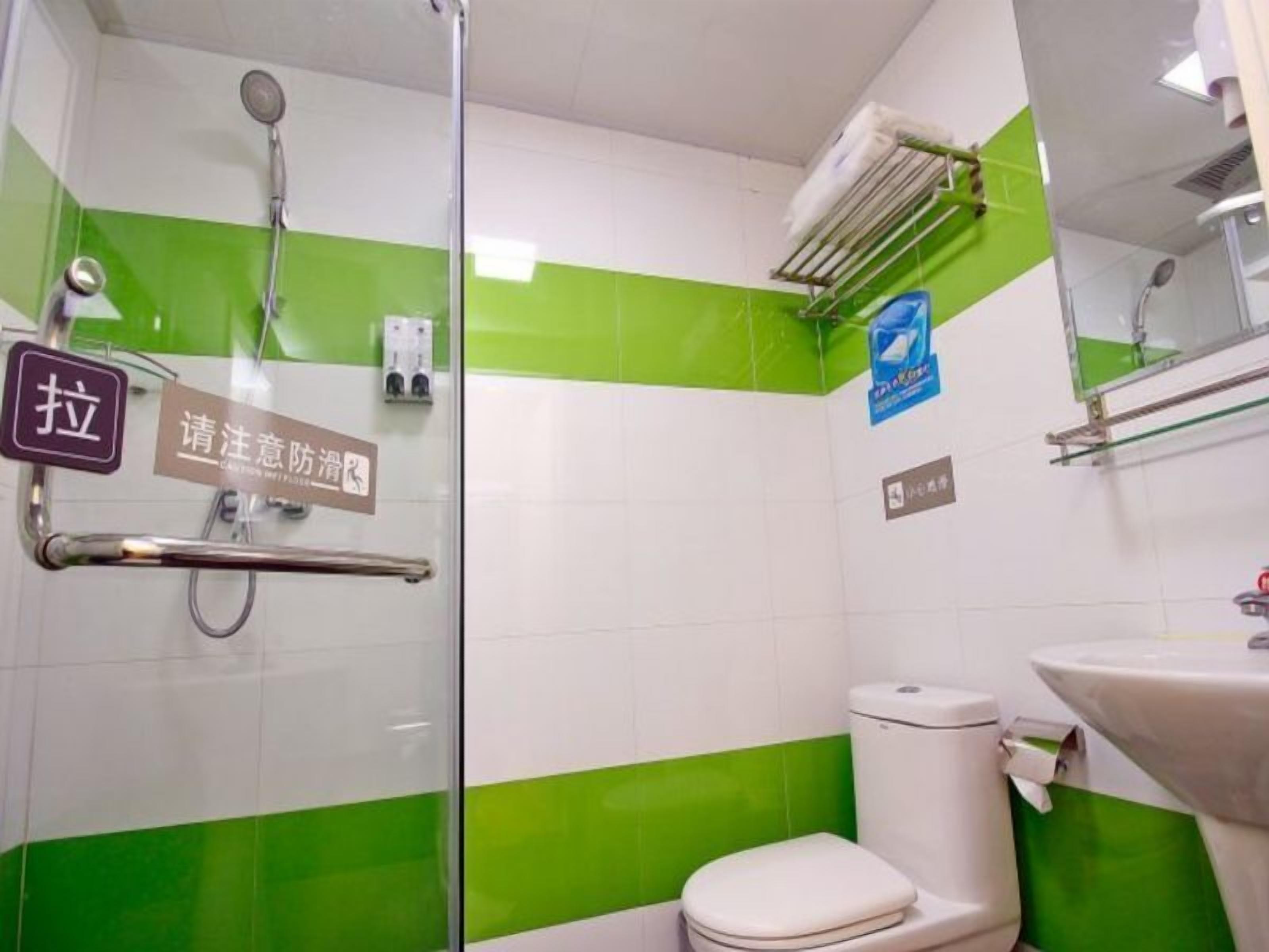 Casa de banho com chuveiro, sanita e lavatório. Apresenta azulejos verdes e brancos, um corrimão e um sinal com caracteres chineses.