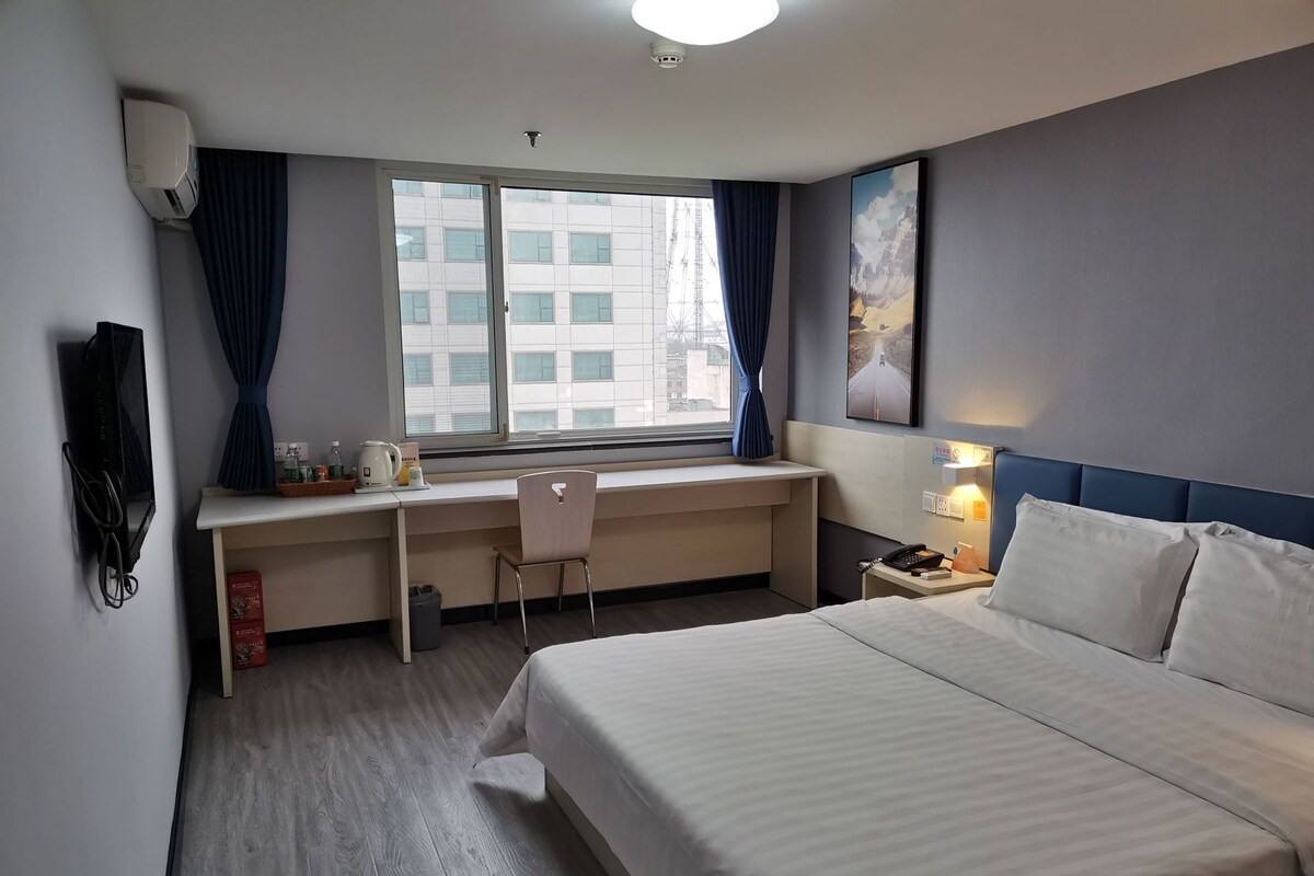 A hotel room with a bed, a desk with a chair, a window with curtains, a wall-mounted TV, and a painting.