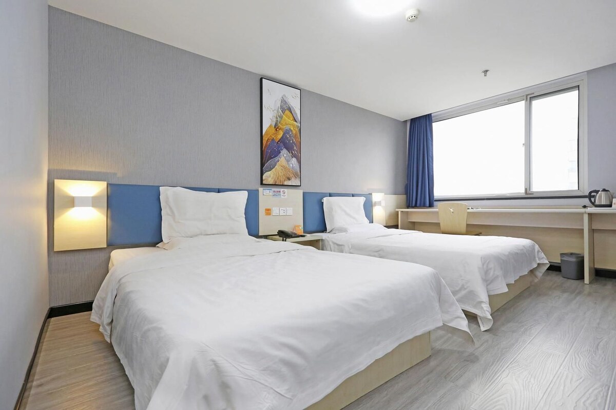 A hotel room with two beds, a desk, a chair, a window, and a wall-mounted artwork.