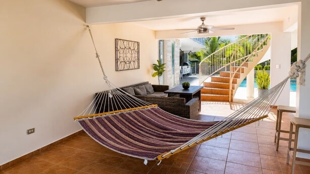 A hammock in a room with a staircase leading to a balcony, a sofa, and a ceiling fan. - Palm's Bohemian House with Private Pool (Aguada)