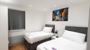 A bedroom with two beds, a window with curtains, a painting on the wall, and a bedside table with a lamp. - Garland Stylish Apartment 2 In Greater London (Upper Belvedere, Kent)