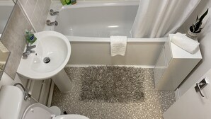 Un baño con lavamanos, inodoro y bañera blancos. Hay una cortina de ducha, una toalla blanca y una pequeña planta en maceta a un lado.