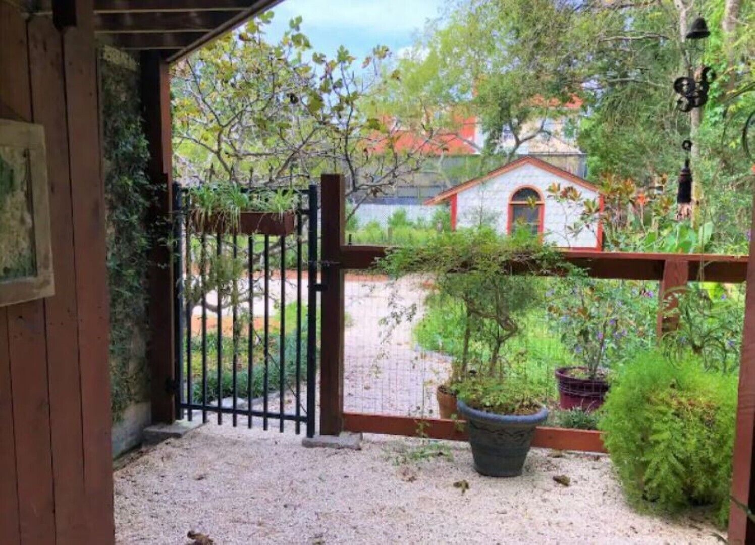 Un cancello di legno che dà su un'area sterrata con vasi di piante e vista su una casa con tetto rosso.
