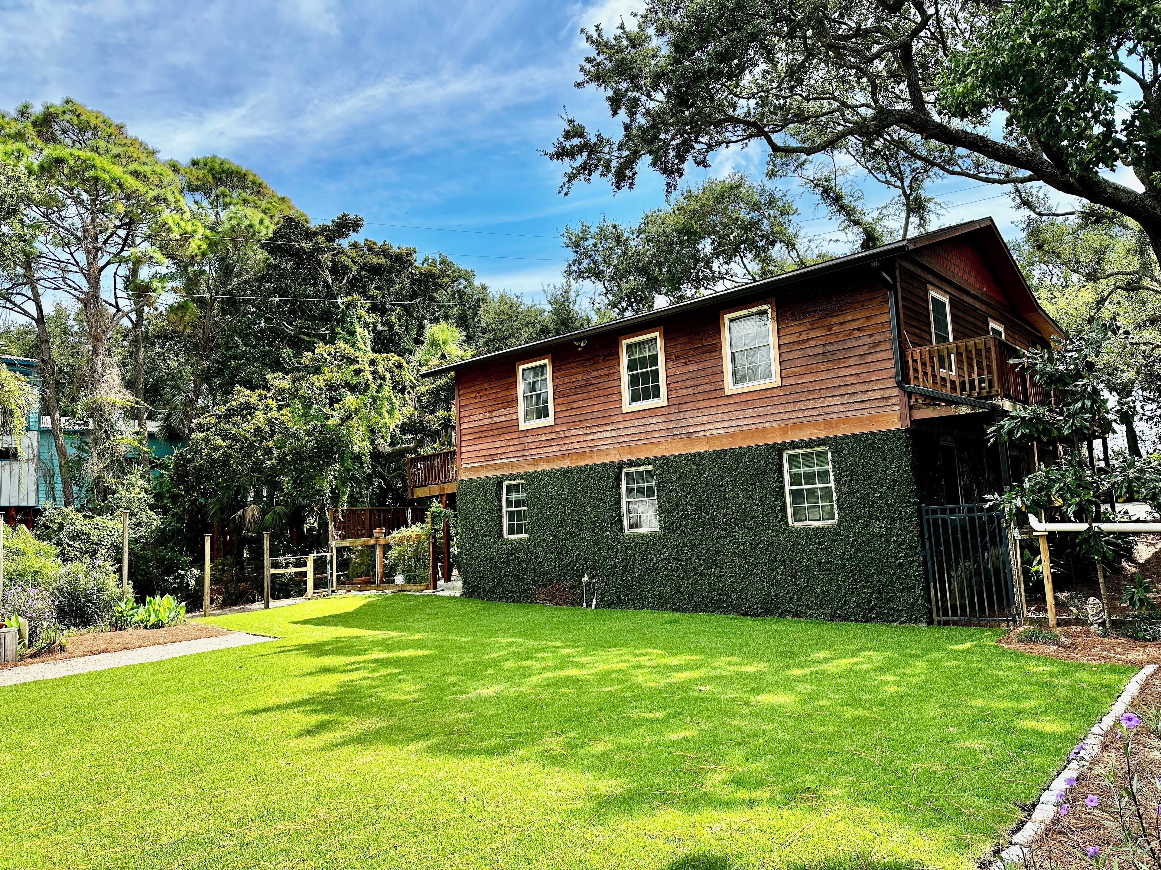 A two-story wooden house with a balcony, surrounded by lush greenery and a well-maintained lawn.