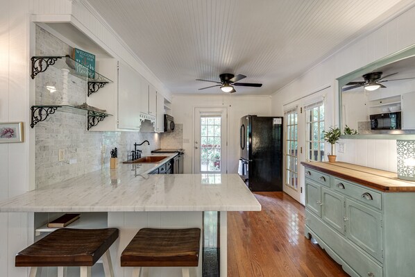 A kitchen with marble countertops, wooden stools, a black refrigerator, and a ceiling fan.