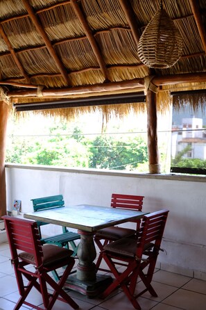 A dining area with a table and chairs, a thatched roof, and a view of greenery outside.
