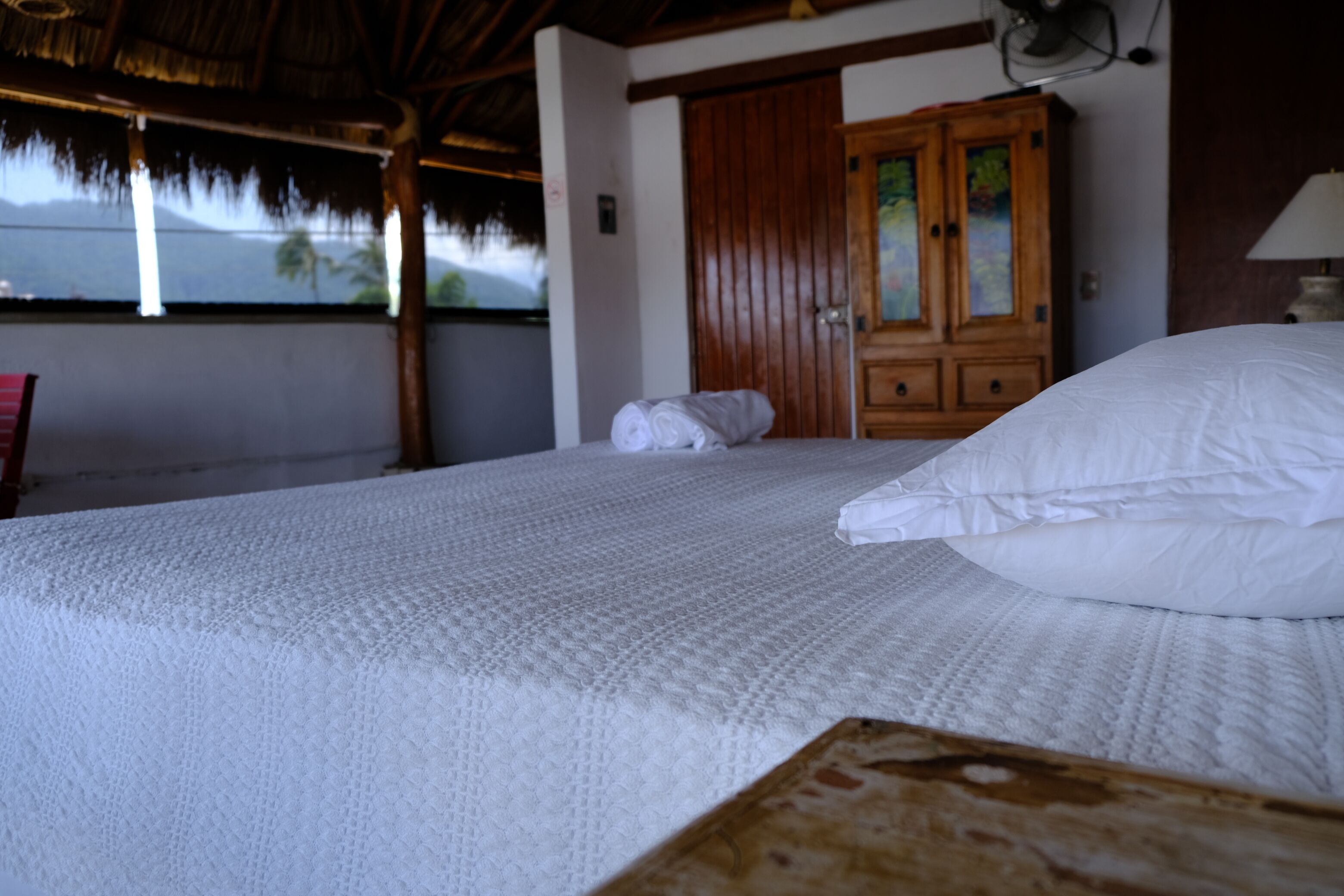 Una cama bien hecha con sábanas blancas, un armario de madera y una ventana con vistas a palmeras.
