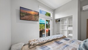 A bedroom with a large bed, a view of the outdoors through a sliding door, a wardrobe, and a mounted TV. - Coastal Escape: 2BR/2B w/King En-suite + Balcony Views, Deck, BBQ (Oceanside)