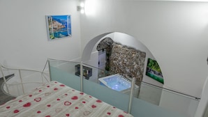 Ein Schlafzimmer mit einem Bett, einer Glaskuppel-Wanne, einer Steinsäule und einem Gemälde einer Küstenlandschaft.